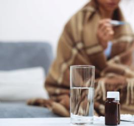 Na pierwszym planie leki i szklanka wody, na drugim kobieta owinięta w koc mierzy sobie temperaturę.