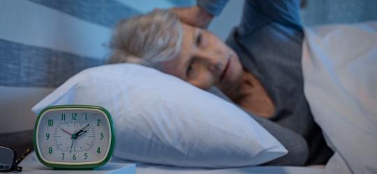 Starsza kobieta leży w łóżku, cierpi na bezsenność. Zegarek pokazuje godzinę 2 w nocy.