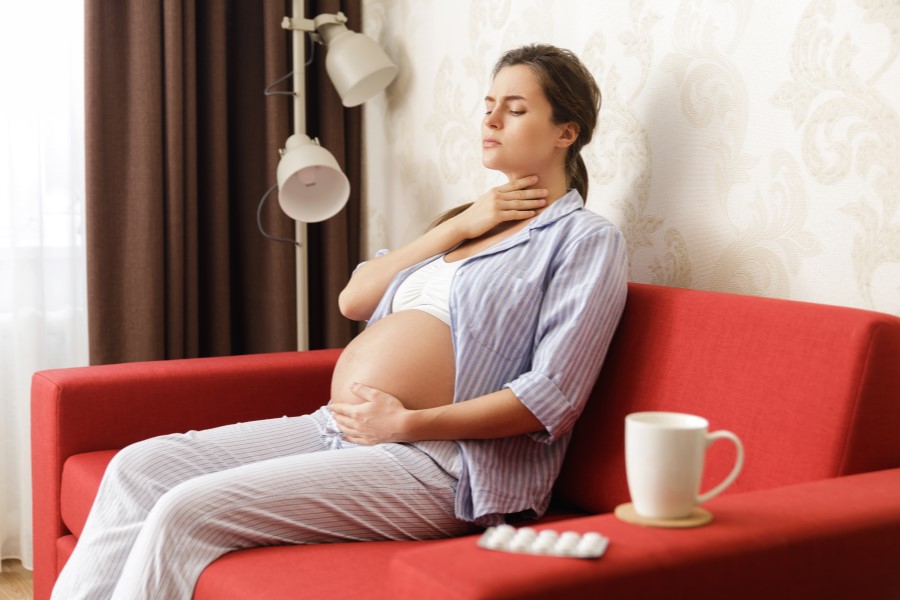 Kobieta w ciąży odczuwa ból gardła, siedzi na czerwonym fotelu, na oparciu leżą leki.