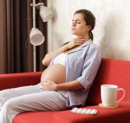 Kobieta w ciąży odczuwa ból gardła, siedzi na czerwonym fotelu, na oparciu leżą leki.