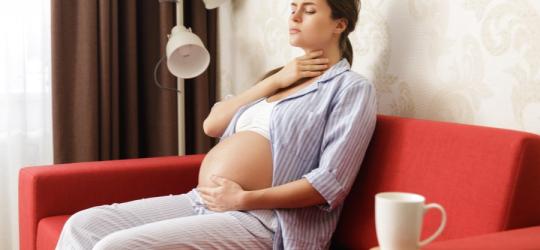 Kobieta w ciąży odczuwa ból gardła, siedzi na czerwonym fotelu, na oparciu leżą leki.