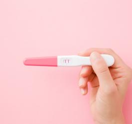 Różowe tło. Kobieca dłoń trzyma test ciążowy z wynikiem pozytywnym.