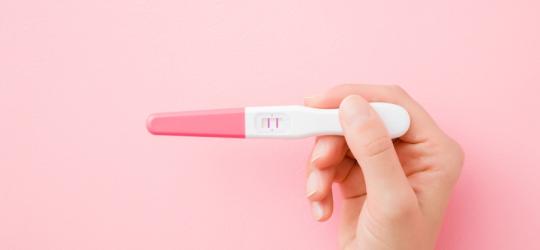 Różowe tło. Kobieca dłoń trzyma test ciążowy z wynikiem pozytywnym.