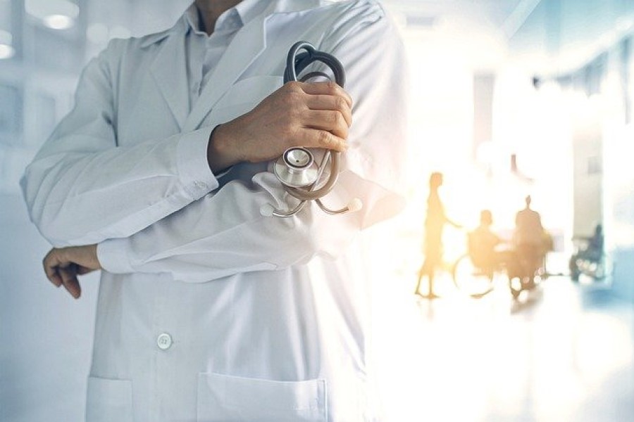 Lekarz w kitlu trzyma w dłoni stetoskop. W tle pielęgniarka i pacjenci w szpitalnym korytarzu.