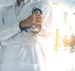 Lekarz w kitlu trzyma w dłoni stetoskop. W tle pielęgniarka i pacjenci w szpitalnym korytarzu.