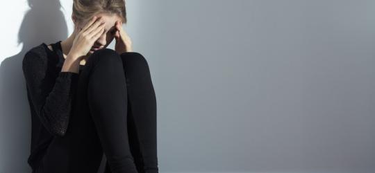 Kobieta siedzi na ziemi, oparta o ścianę, jest zdenerwowana i smutna.