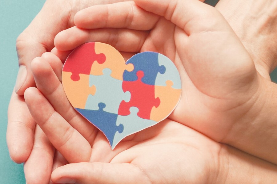 Ręce dziecka trzymane przez ręce rodzica. W złączonych dłoniach widoczne serce z puzzli.