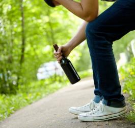 Mężczyzna siedzący na ławce w parku, trzyma w dłoni butelkę z alkoholem.