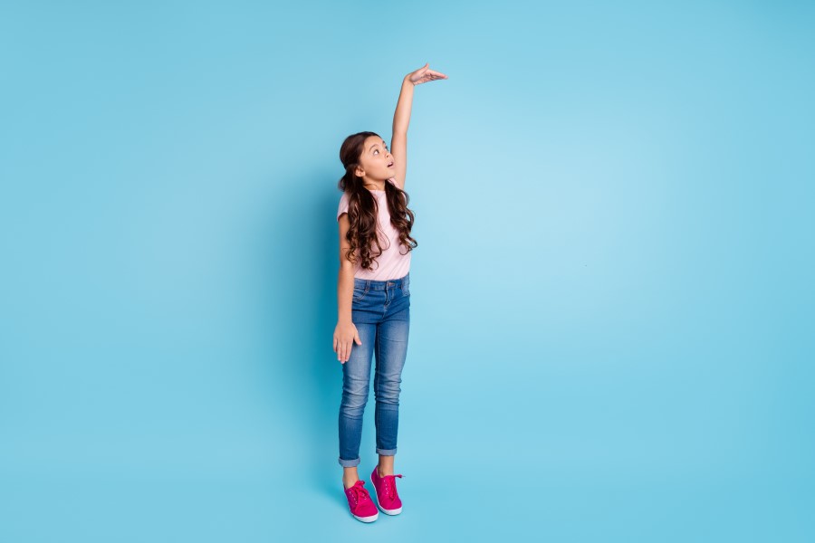 Dziewczynka stoi na palcach, unosi rękę do góry wskazując wysoki wzrost.