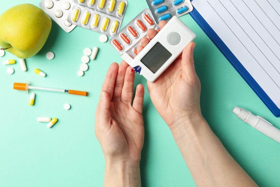Pacjent wykonuje pomiar cukru glukometrem. W tle leżą leki, notatnik, pen, jabłko.