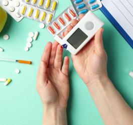 Pacjent wykonuje pomiar cukru glukometrem. W tle leżą leki, notatnik, pen, jabłko.