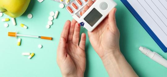 Pacjent wykonuje pomiar cukru glukometrem. W tle leżą leki, notatnik, pen, jabłko.