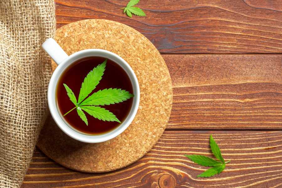 Biały kubek na drewnianym blacie i korkowej podstawce. W kubku zaparzona herbata konopna.