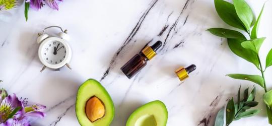 Kosmetyki, awokado, zegarek i kwiaty na marmurowym blacie.