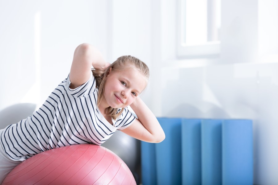 Uśmiechnięta dziewczynka na piłce gimnastycznej wykonuje ćwiczenia na kifozę.