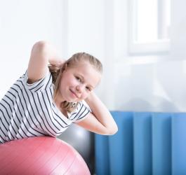 Uśmiechnięta dziewczynka na piłce gimnastycznej wykonuje ćwiczenia na kifozę.