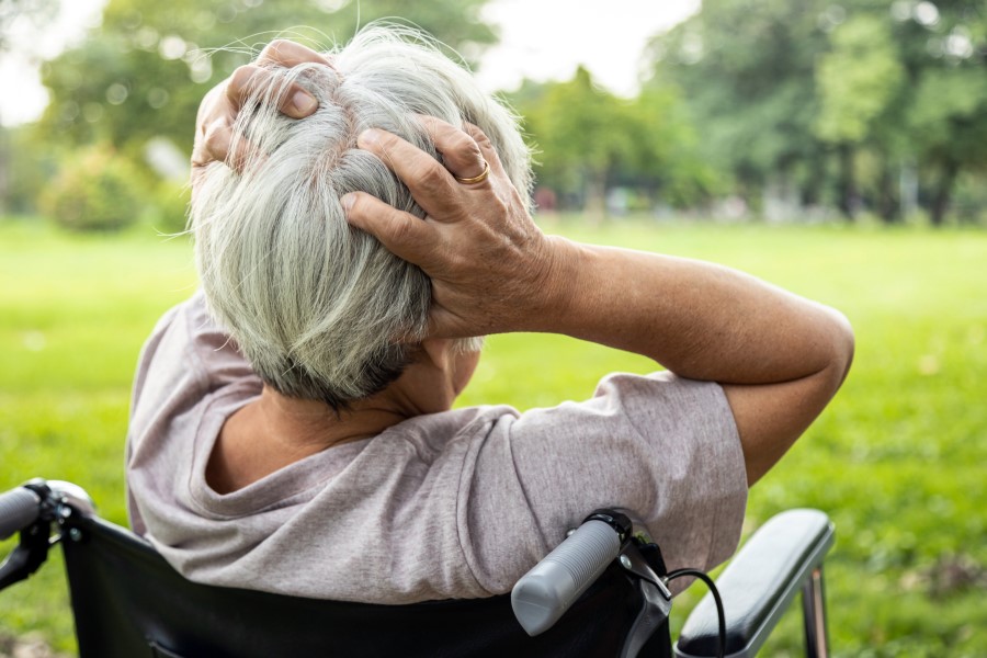 Starsza kobieta siedzi na wózku inwalidzkim, trzyma się dłońmi za głowę.