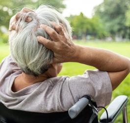 Starsza kobieta siedzi na wózku inwalidzkim, trzyma się dłońmi za głowę.