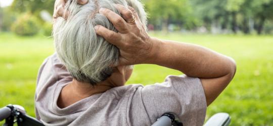 Starsza kobieta siedzi na wózku inwalidzkim, trzyma się dłońmi za głowę.