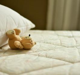 Pluszowy miś leży na materacu łóżka, oparty o poduszkę.