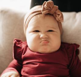 Niemowlę, dziewczynka robi zabawny grymas. Ubrana jest w bordową bluzkę i różową opaskę z kokardą.