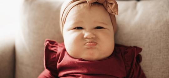 Niemowlę, dziewczynka robi zabawny grymas. Ubrana jest w bordową bluzkę i różową opaskę z kokardą.