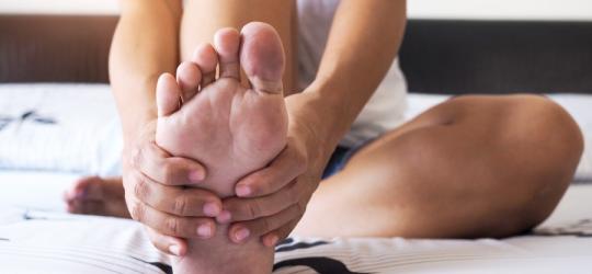 Osoba trzyma się dłońmi za stopę, doskwierają jej bolesne nagniotki.