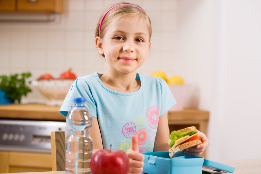 Dziewczynka przygotowuje swój lunchbox do szkoły, zawierający kanapkę, jabłko i butelkę wody.