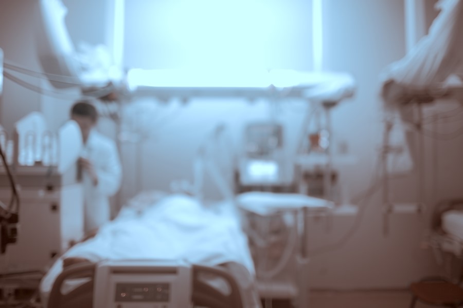 Rozmazane zdjęcie sali szpitalnej z pacjentem na łóżku i lekarzem przy aparaturze medycznej.