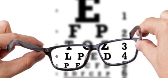Pacjent trzyma okularu korekcyjne, w tle tablica okulistyczna.