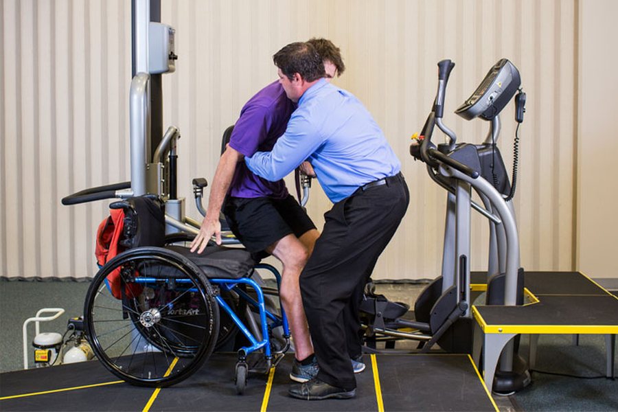 Rehabilitant pomaga pacjentowi wstać z wózka inwalidzkiego przy użyciu sprzętu do rehabilitacji.