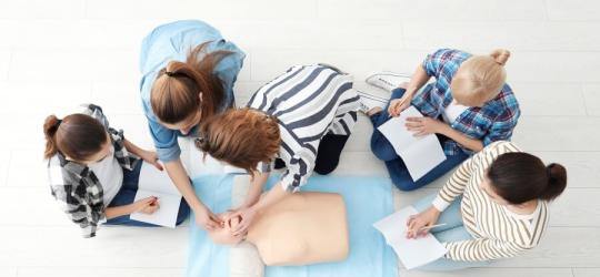 Grupa 5 osób podczas kursu resuscytacji krążeniowo-oddechowej. Robią notatki i ćwiczą na fantomie.