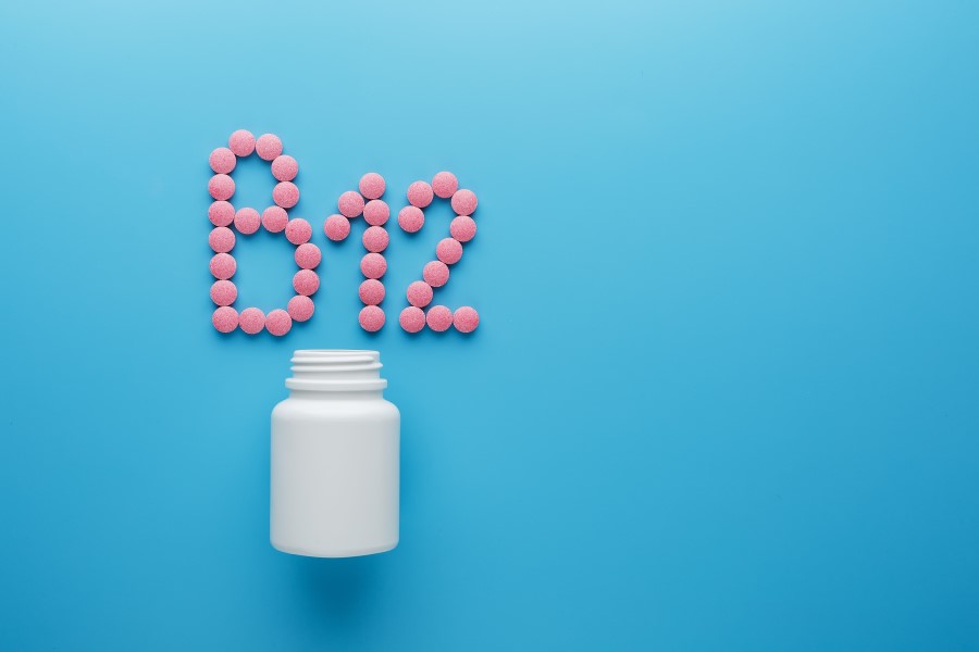 Różowe tabletki na niebieskim tle ułożone w napis B12, obok biała buteleczka.