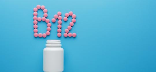 Różowe tabletki na niebieskim tle ułożone w napis B12, obok biała buteleczka.