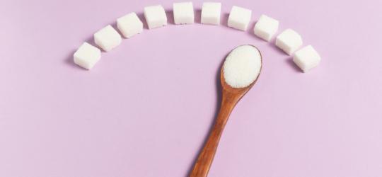 Różowe tło, na którym leżą kostki cukru i drewniana łyżeczka wypełniona cukrem.