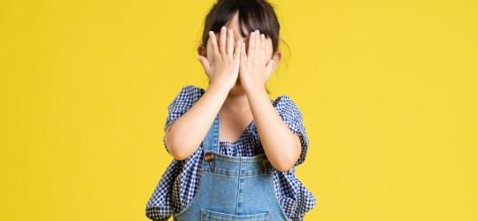 Dziewczynka zasłania oczy i twarz dłońmi.