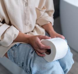 Kobieta siedzi na toalecie, trzyma w dłoniach papier toaletowy. Dokuczają jej zaparcia.