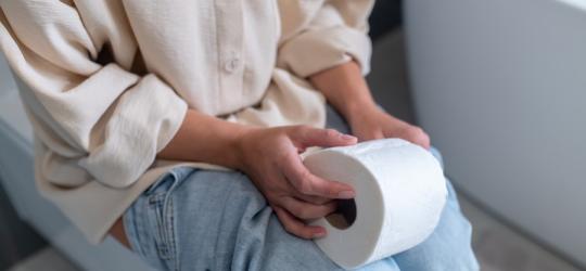 Kobieta siedzi na toalecie, trzyma w dłoniach papier toaletowy. Dokuczają jej zaparcia.