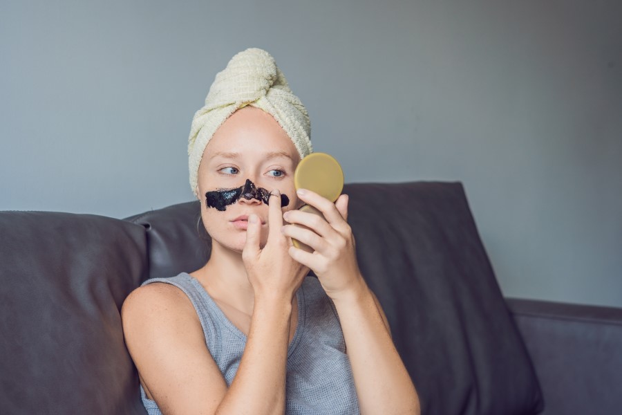 Kobieta nakłada na nos i policzki maseczkę na zaskórniki, przegląda się w lusterku.