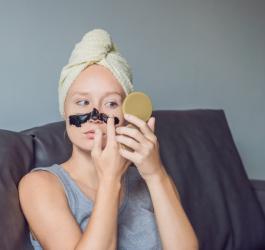 Kobieta nakłada na nos i policzki maseczkę na zaskórniki, przegląda się w lusterku.