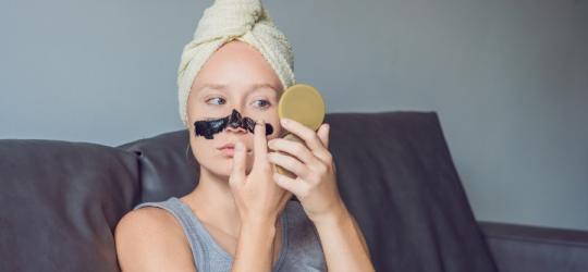 Kobieta nakłada na nos i policzki maseczkę na zaskórniki, przegląda się w lusterku.