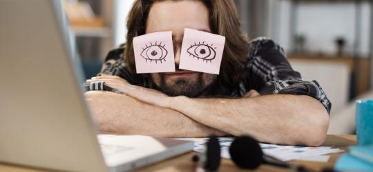 Przemęczony mężczyzna przysypia na biurku przed otwartym laptopem.