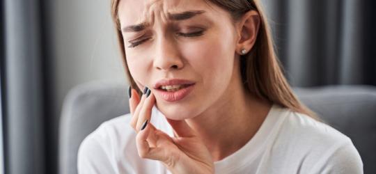 Kobieta trzyma się za żuchwę. Widać, że odczuwa silny ból, co może być spowodowane zgorzelą zęba.
